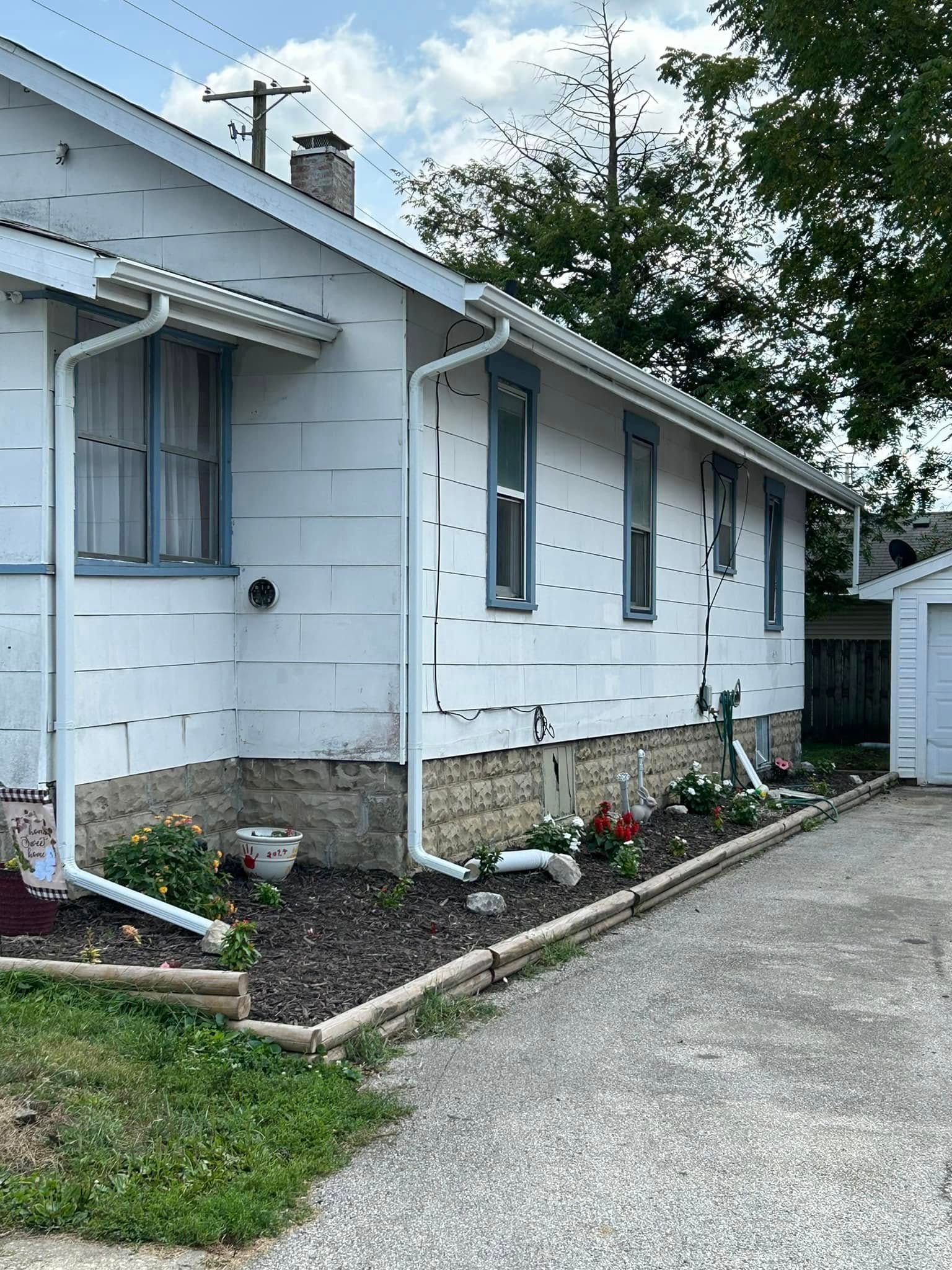 White house with blue-trimmed windows, gutters, and a flower bed. Gravel driveway and a garage.
