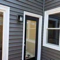 Exterior of a house with gray siding, white trim around a glass door and window. A light is mounted beside the door.