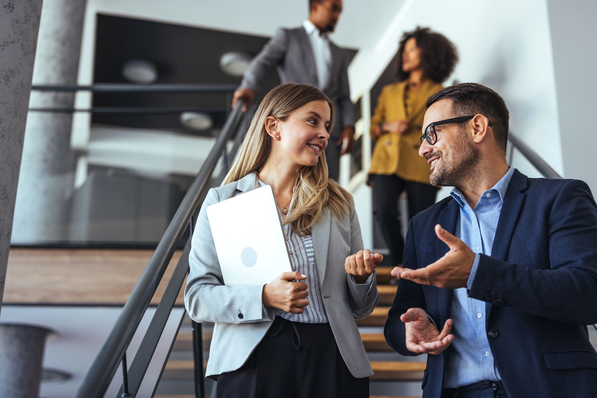Two coworkers talking on an office staircase, smiling and holding a laptop and phone
