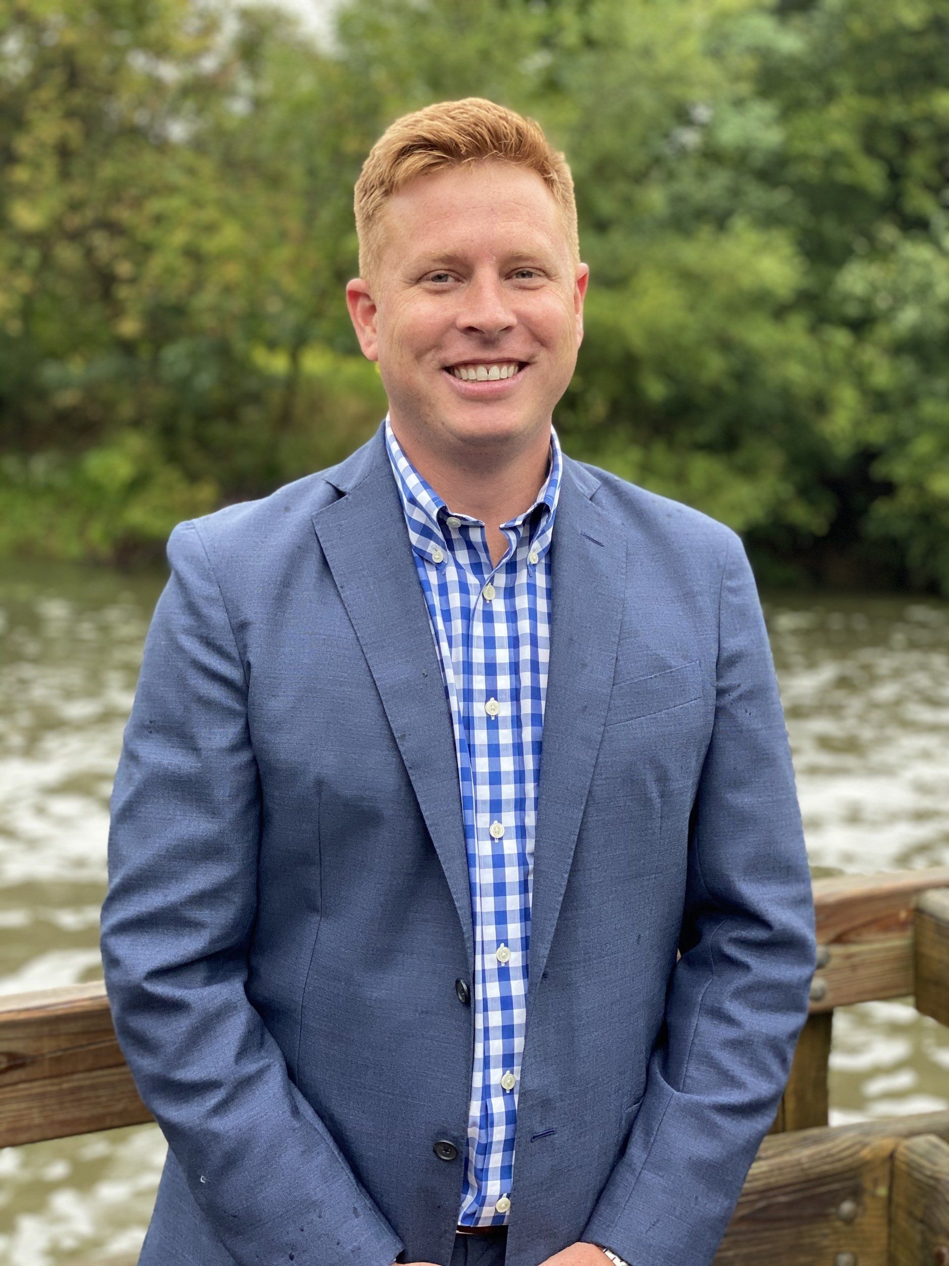 A man in a suit and plaid shirt is standing on a bridge overlooking a body of water.