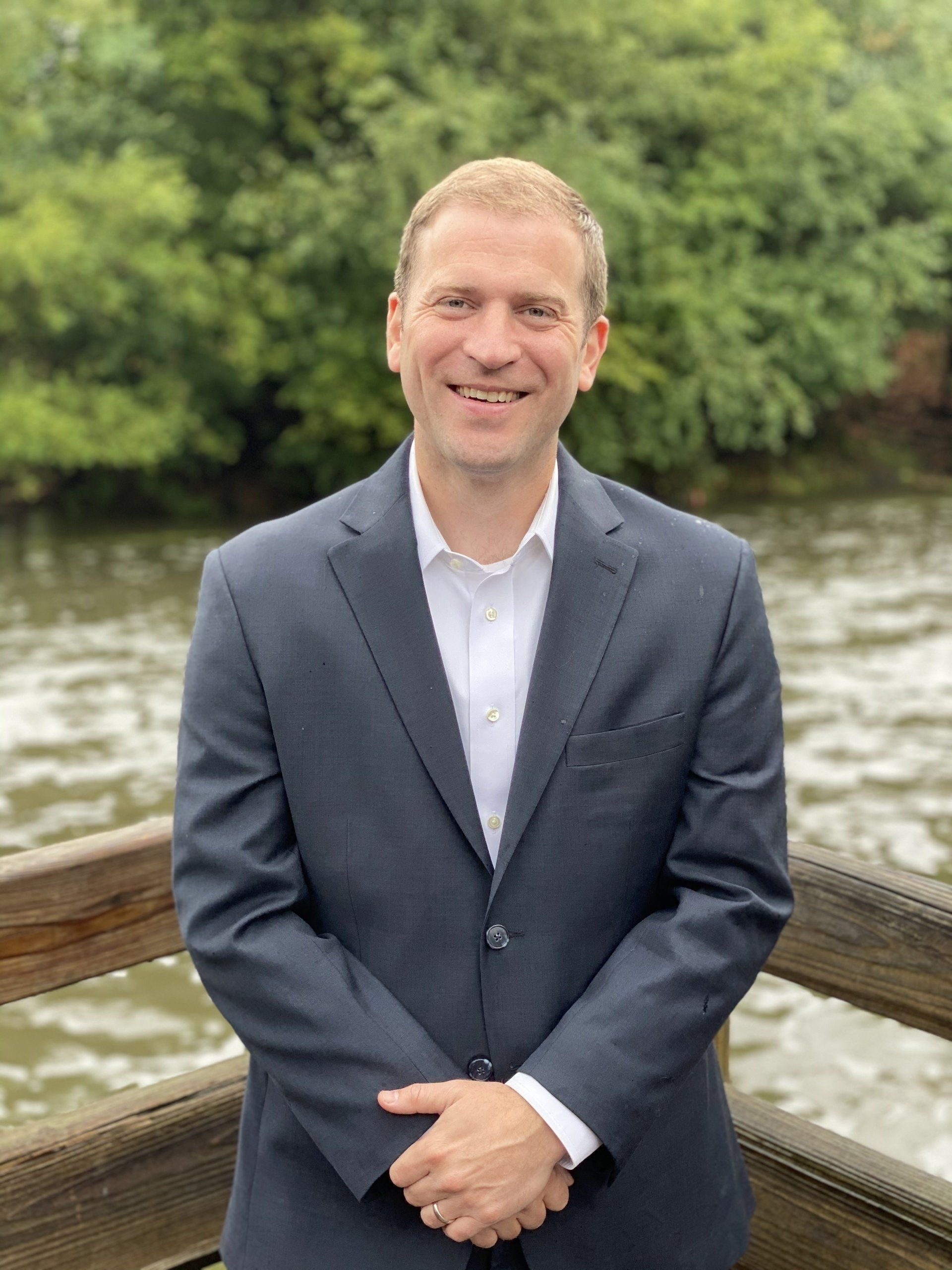 A man in a suit and white shirt is standing on a bridge overlooking a body of water.