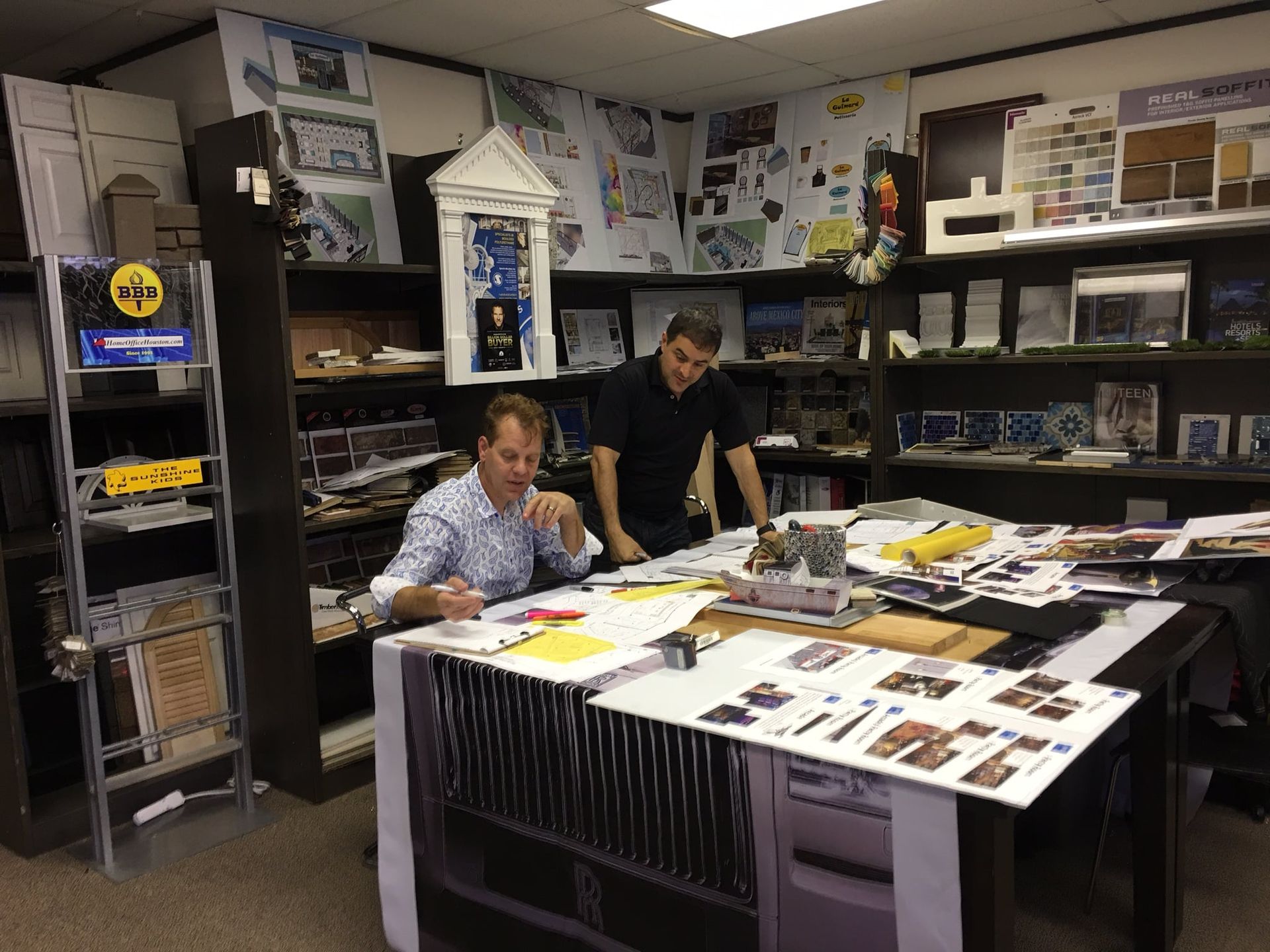 Two men review documents at a desk in a cluttered office with architectural models and drawings.