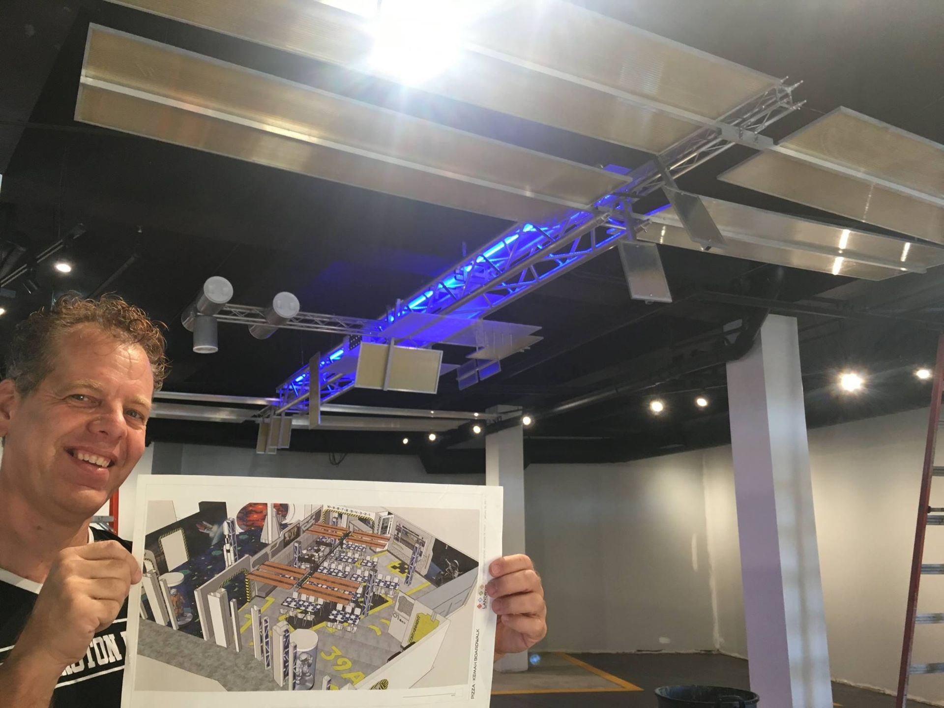 Man smiles, holding architectural drawing in a large room with ceiling lights and truss; construction underway.