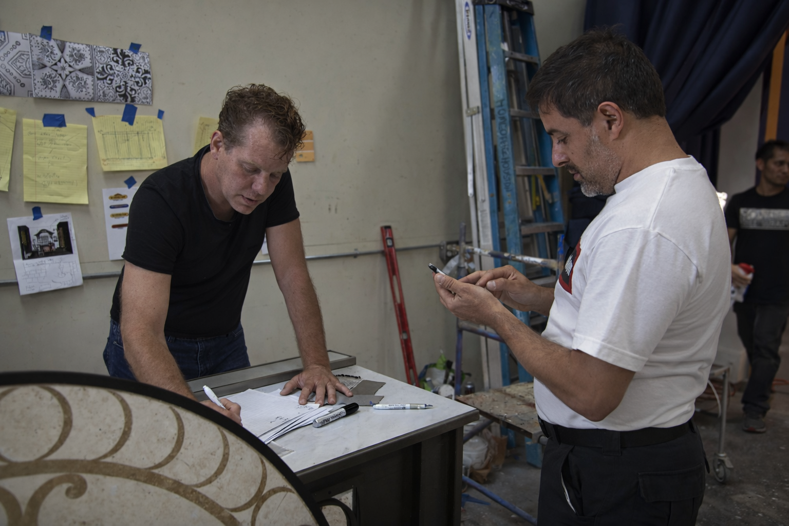 Two men collaborating over a design at a desk, papers and sketches visible. One holds a small item, the other writes.