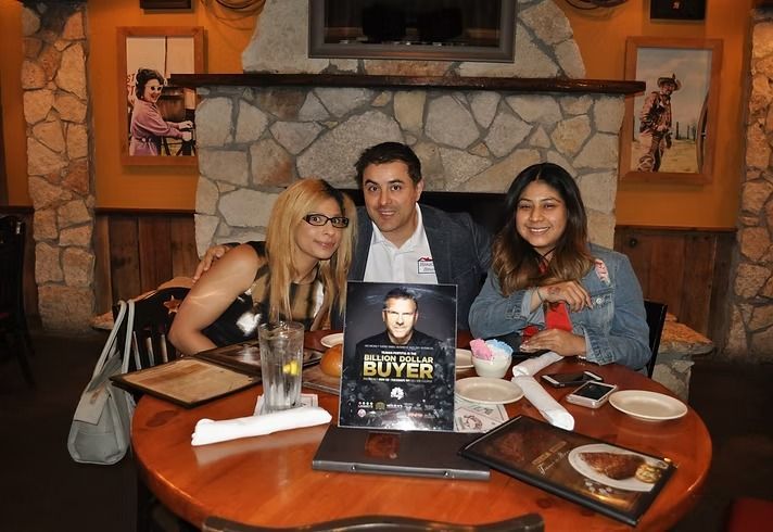 Three people sit at a round wooden table in a restaurant with a stone fireplace, posing with a flyer for a real estate event.