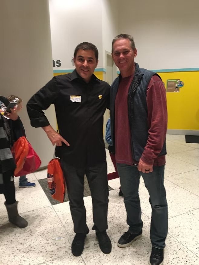 Two men smiling for a photo in a hallway with yellow walls. One wears a black shirt, the other a red shirt and dark vest.
