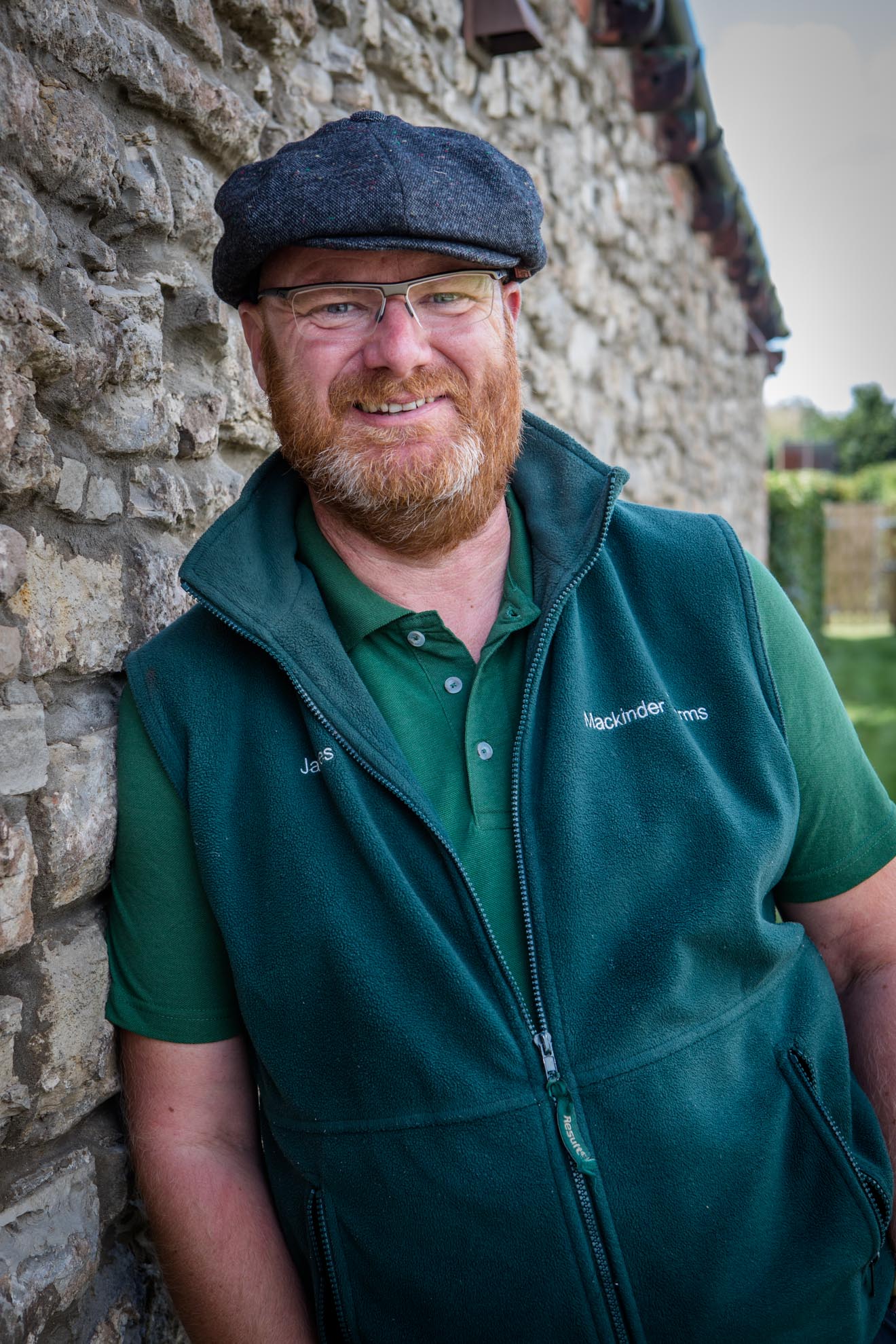 A man with a beard wearing a hat and glasses is standing in front of a stone wall.
