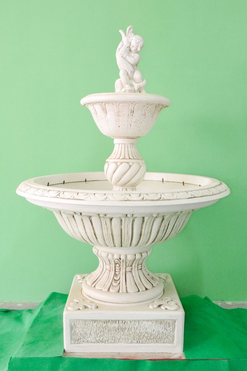 Fontana decorativa in pietra bianco a due livelli con una parte superiore scultorea, posta su uno sfondo verde semplice.