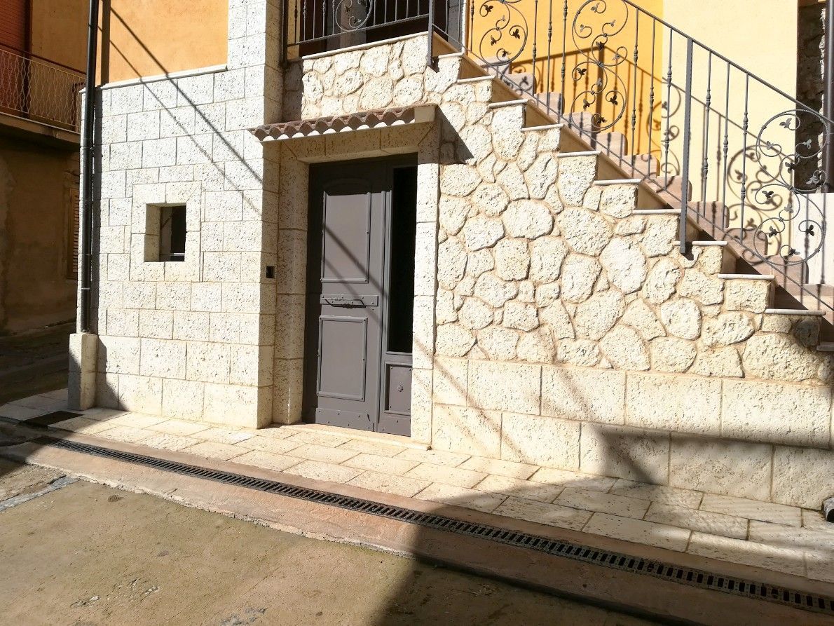 L'ingresso di un edificio con pareti in pietra, una porta grigia, una finestra e scale esterne con una ringhiera in ferro.