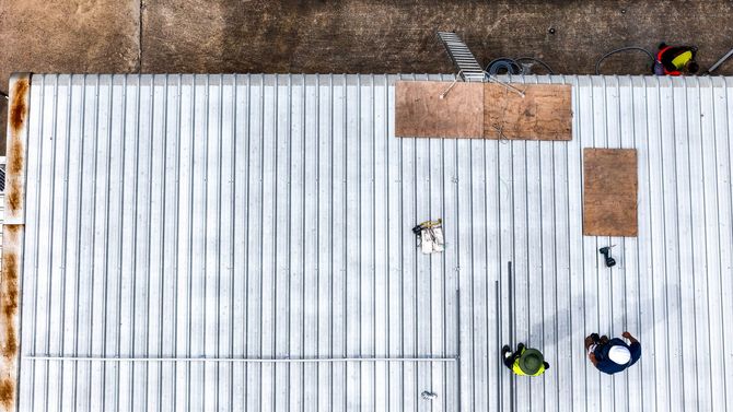 Aerial view of workers on a corrugated metal roof with plywood sheets and a ladder.
