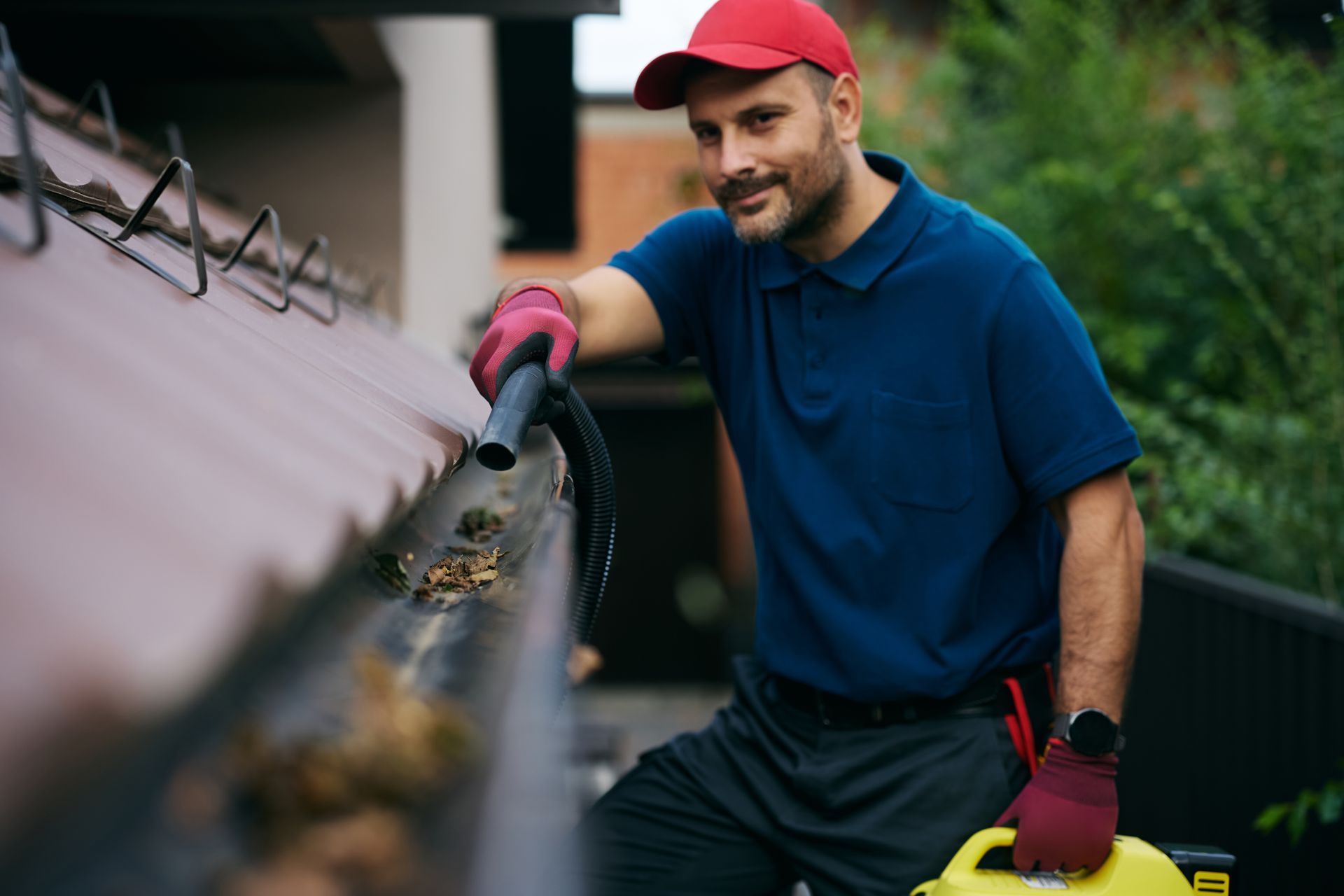 A person wearing a blue polo, red cap, and pink gloves cleans debris from a house gutter using a vacuum nozzle.