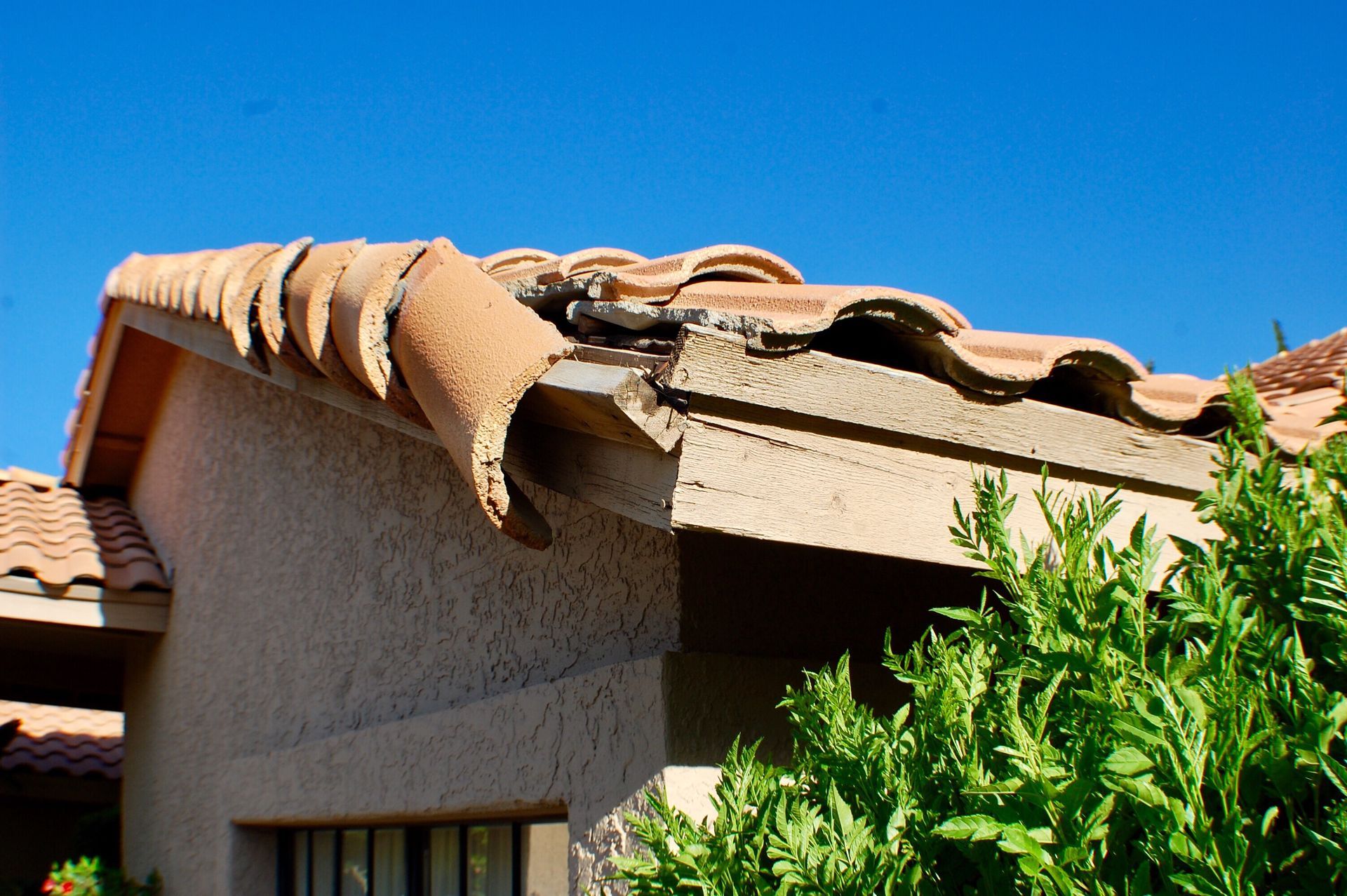 Damaged, detached clay roof tiles hanging over the edge of a house exterior against a bright blue sky.