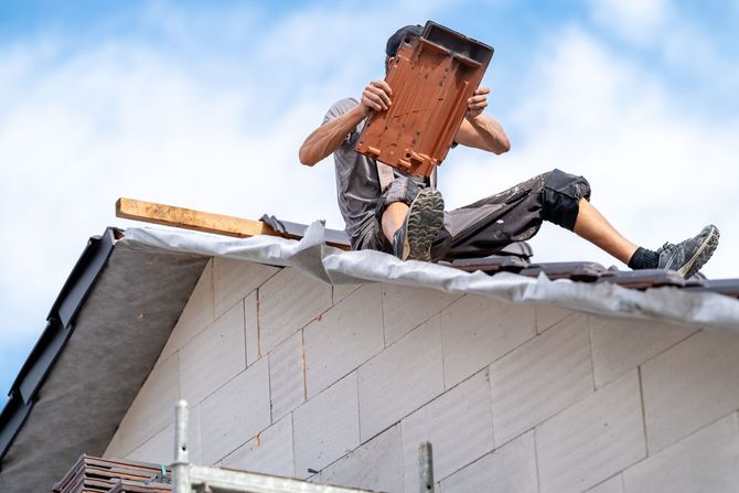 A construction worker in a grey uniform sits on a roof, carefully installing a single roof tile against a blue sky.