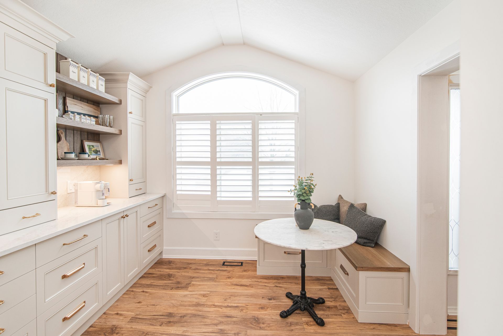 A kitchen with white cabinets , wooden floors , a table and a window.