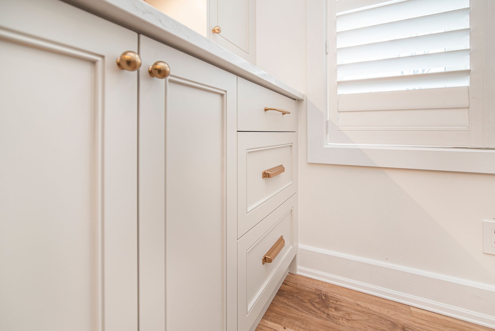 A kitchen with white cabinets and drawers and a window with shutters.