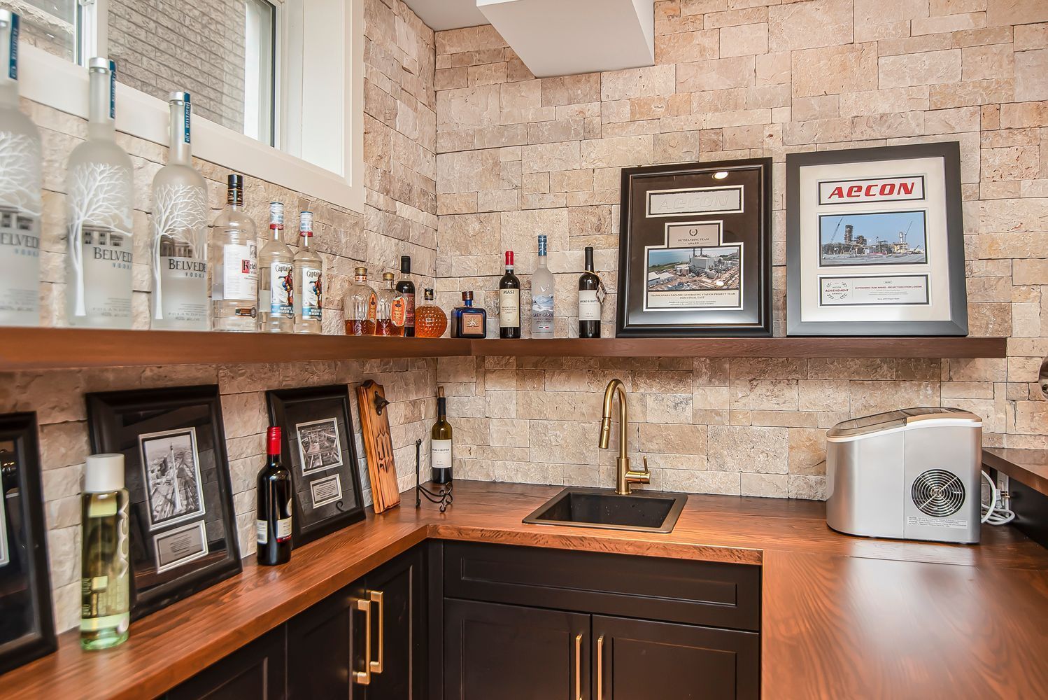 A kitchen with a sink , shelves , bottles of alcohol and pictures on the wall.