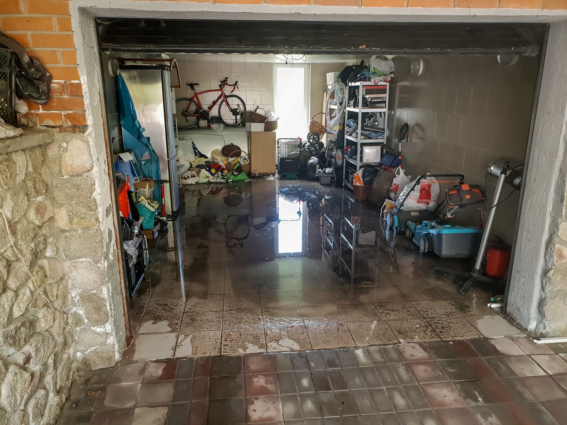 Flooded garage with muddy water, bicycles, shelves, and tools inside open doorway