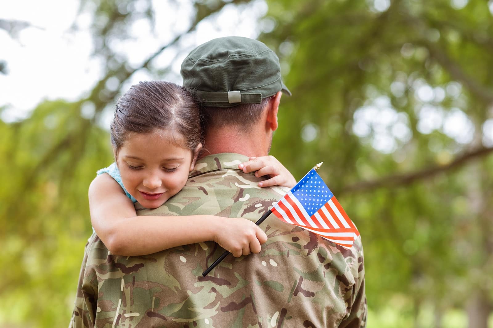 Soldier in camouflage carrying a smiling child with a small American flag on his shoulder outdoors