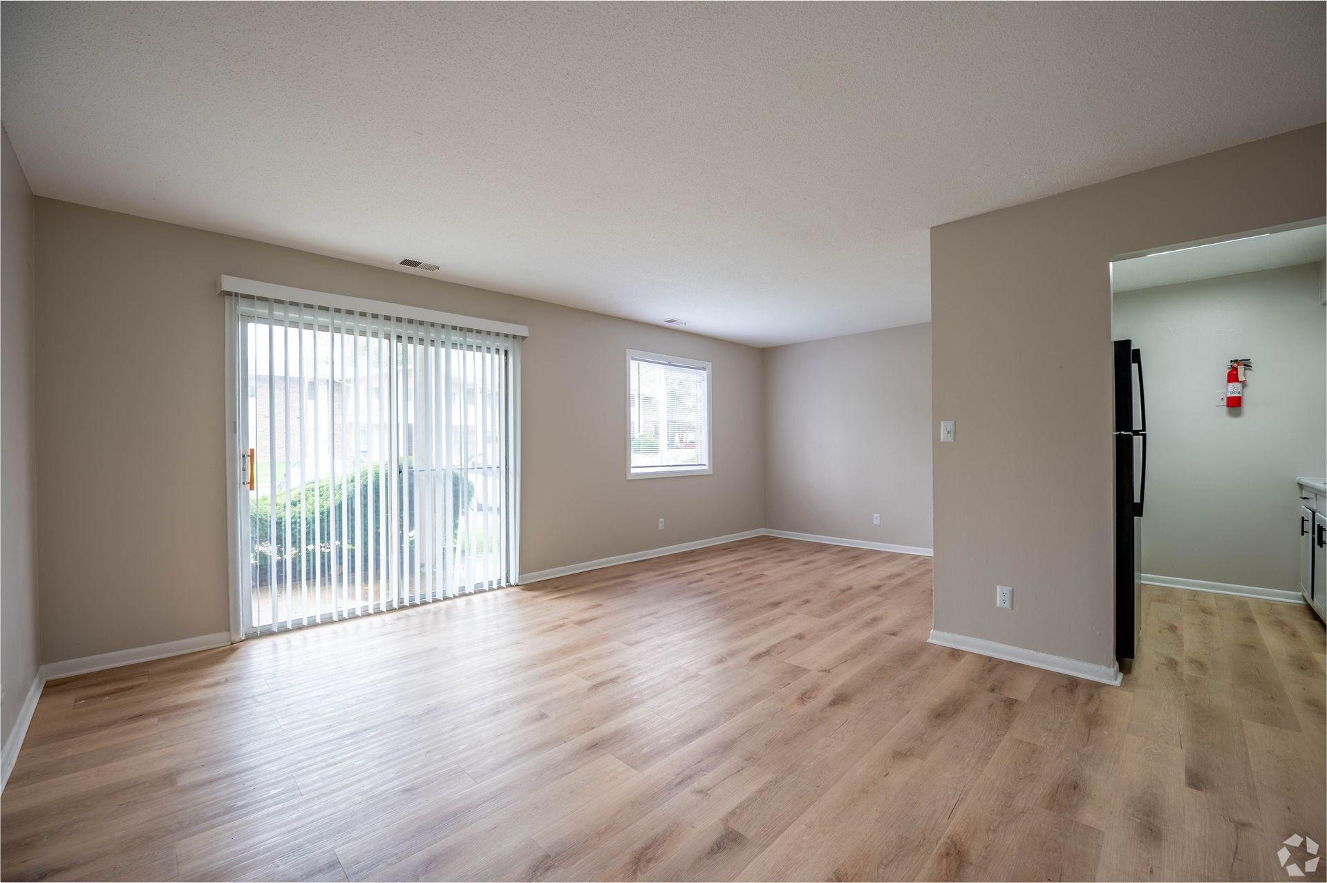 Empty living room with wood-look floors, sliding glass door, window, and access to the kitchen.
