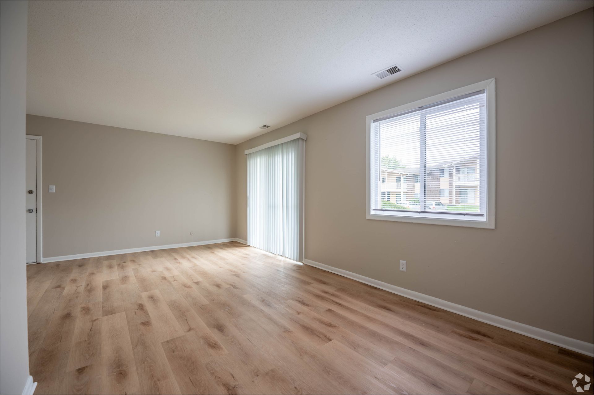 Empty living room with wood-look floors, sliding glass door, and a window with blinds.
