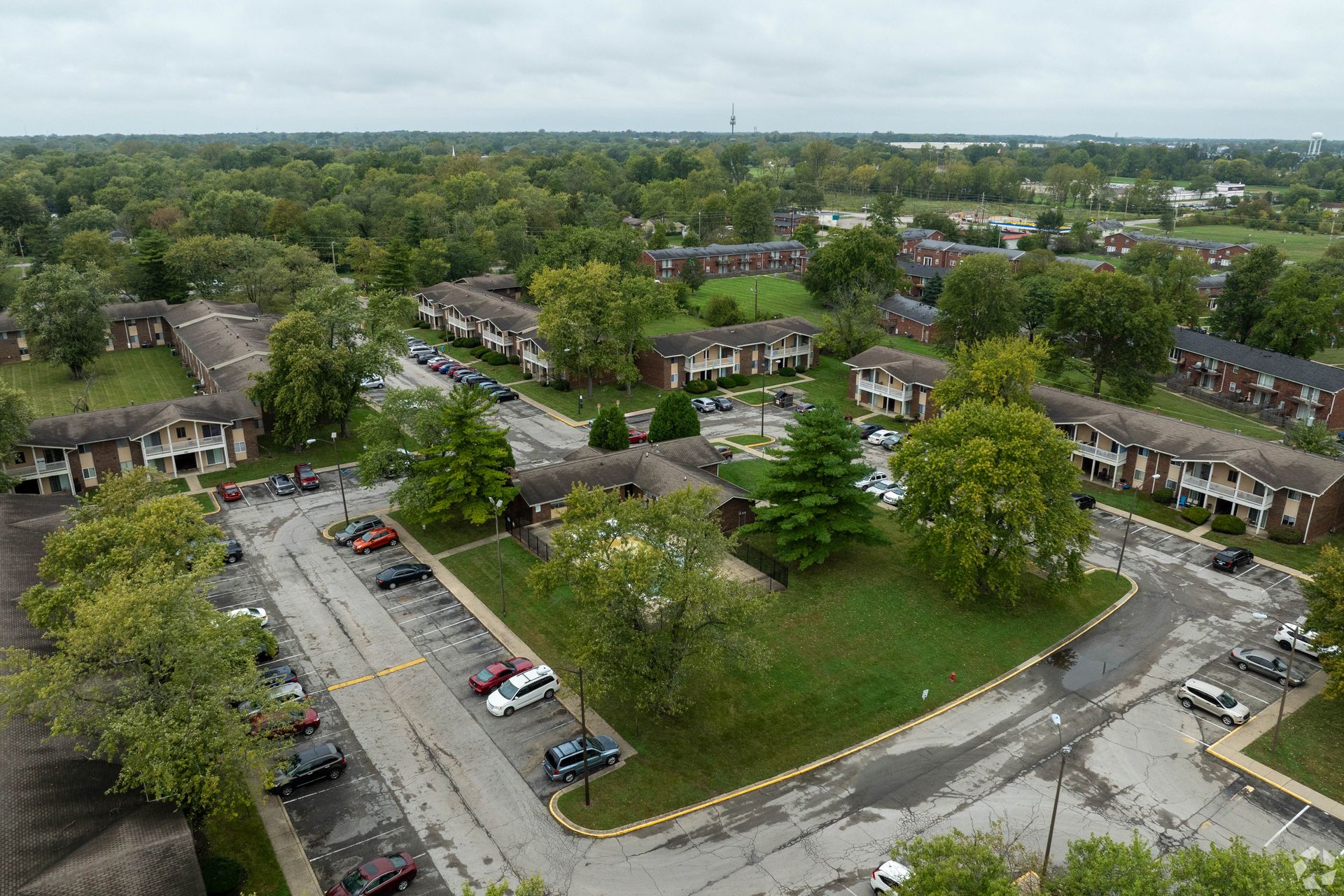 Aerial view of an apartment complex surrounded by trees and greenery, with parked cars in a parking lot.