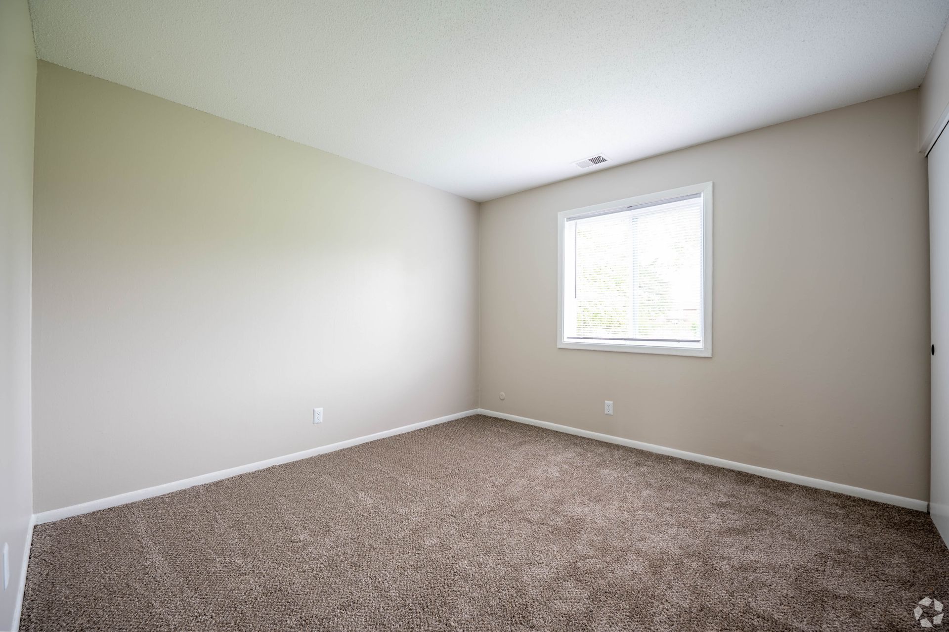 Empty bedroom with beige walls, brown carpet, and a window.