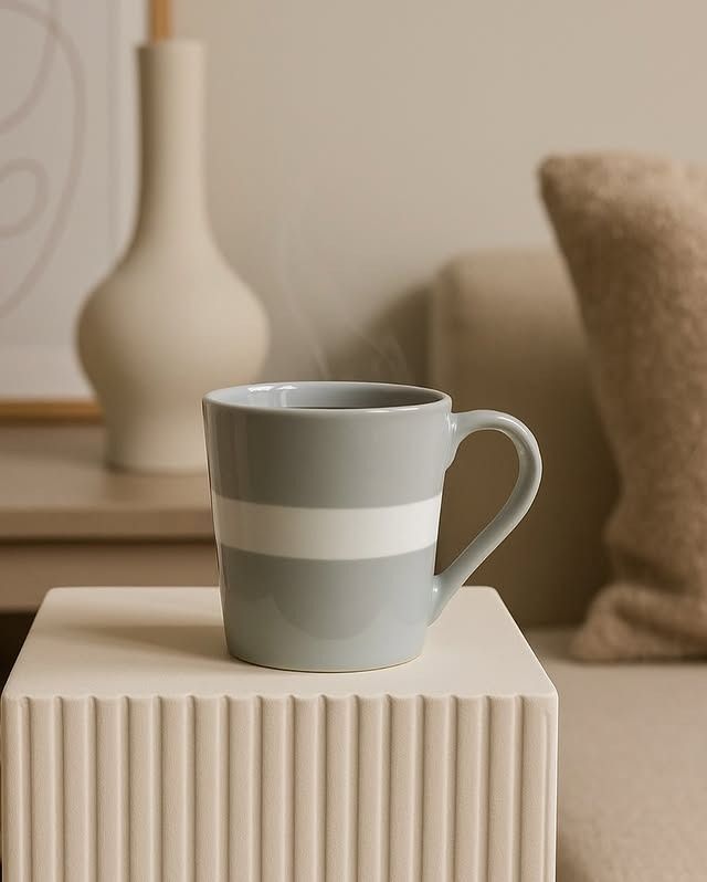 Taza gris con raya blanca, humeante, sobre un bloque de crema, cerca de un jarrón y una almohada.
