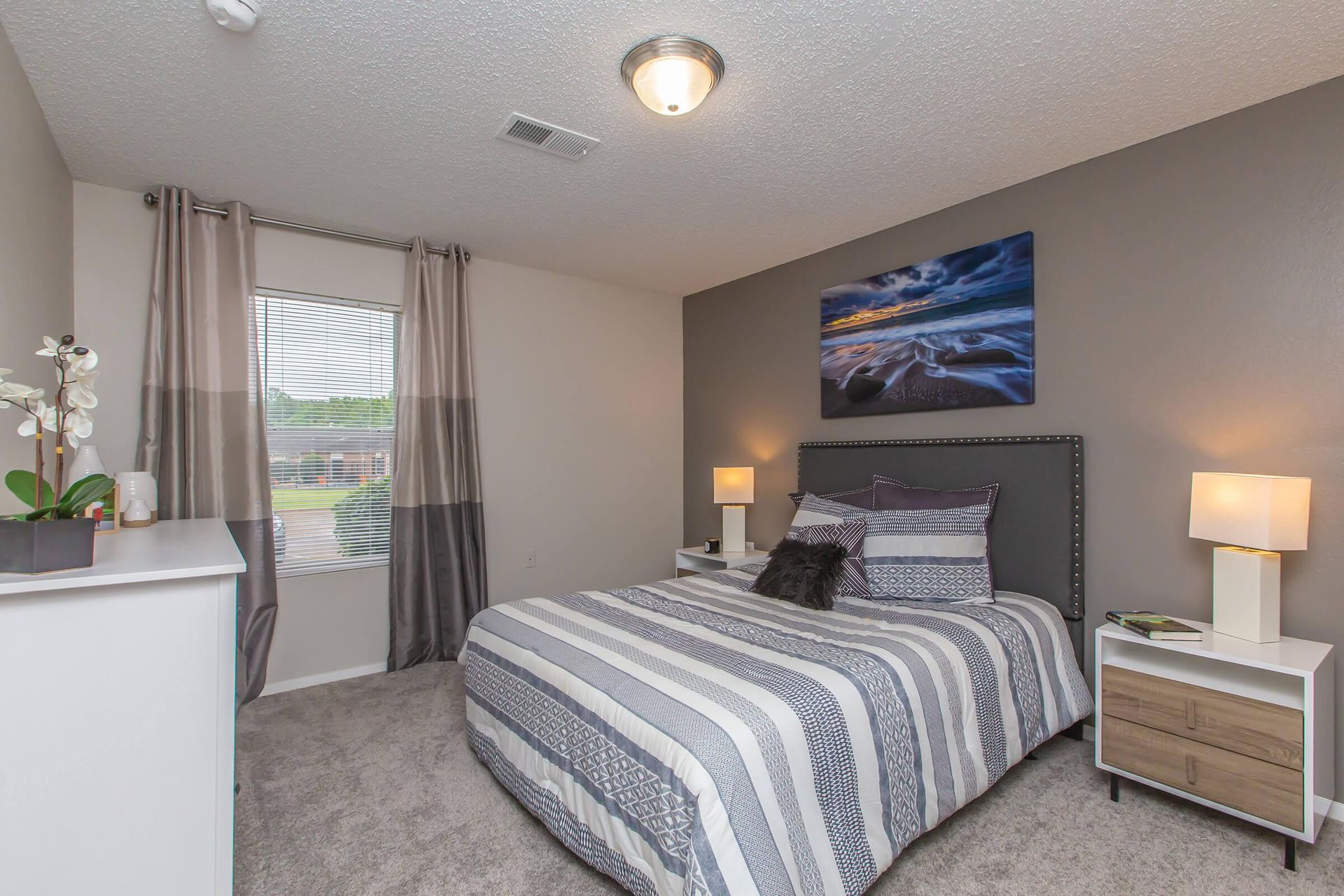 Bedroom with bed, bedside tables, art, window, and dresser. Gray walls, striped bedding, and carpet.