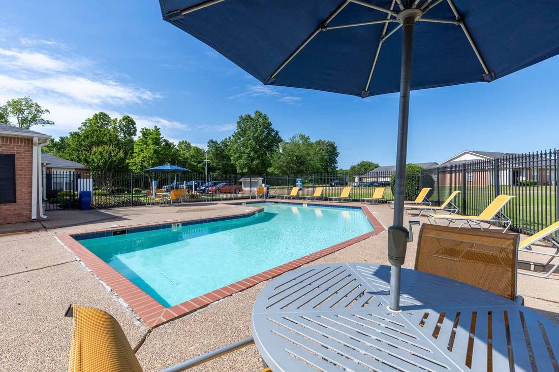 Poolside seating with a table, orange chairs, and blue umbrella under a blue sky.