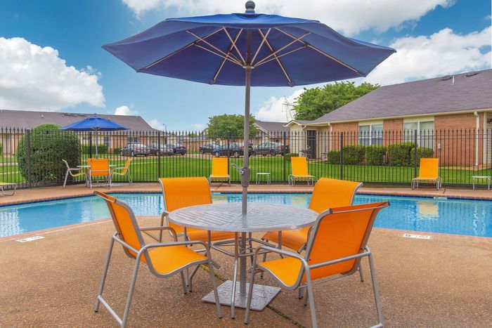 Poolside seating with a table, orange chairs, and blue umbrella under a blue sky.