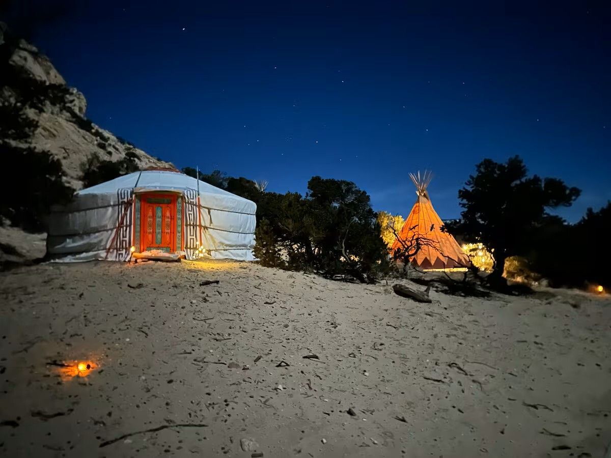 A yurt and teepee illuminated at night on sandy terrain, under a starlit sky.