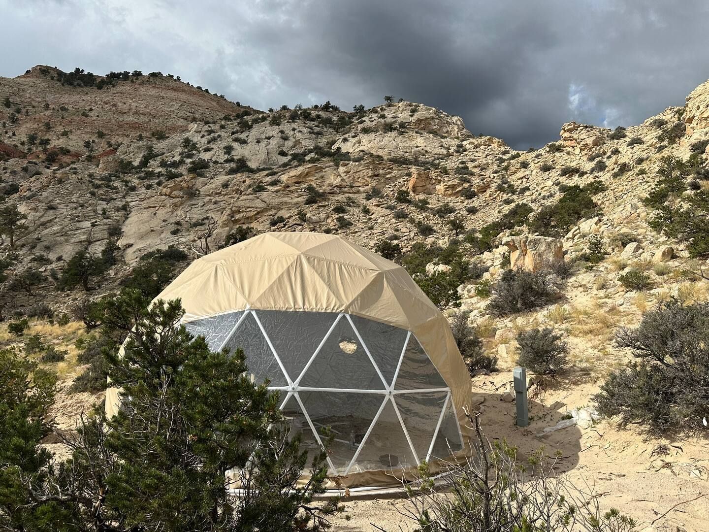 Dome-shaped structure nestled in a rocky, desert landscape. Beige roof, clear lower panels, overcast sky.