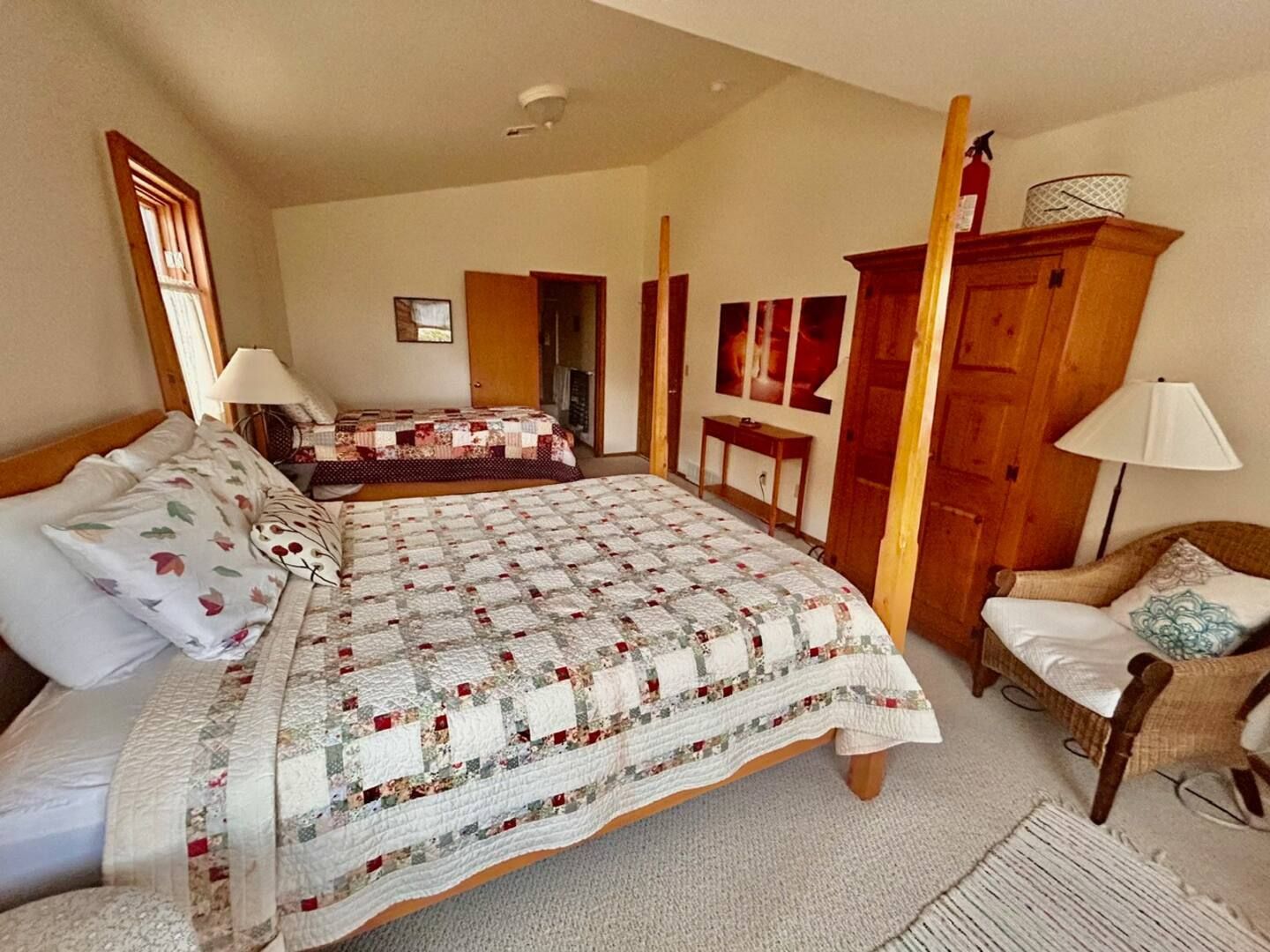 Bedroom with two beds, wooden wardrobe, lamp, and wicker chair, with a white and tan quilt on a bed.