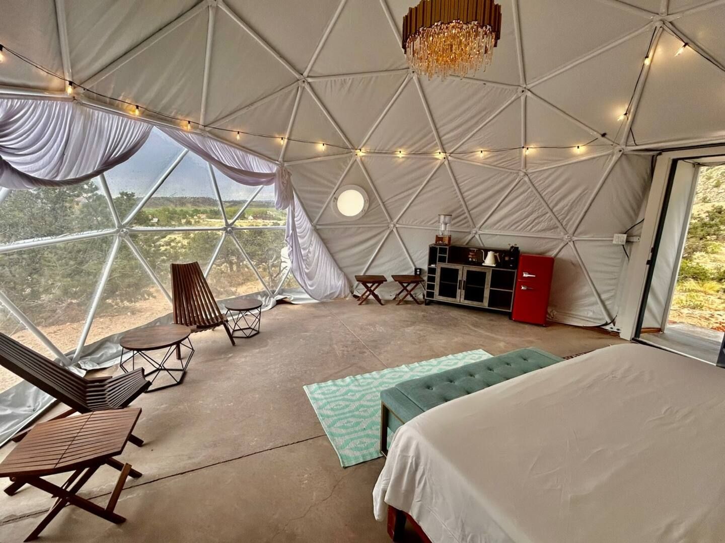Interior of a geodesic dome: bedroom with seating, bed, rug, small fridge, and string lights. 
