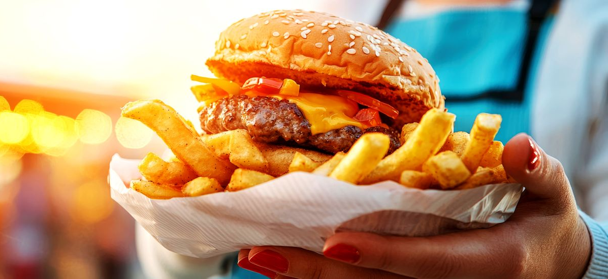 Person holding a burger and fries in a paper holder. The burger has cheese, toppings, and a sesame seed bun.
