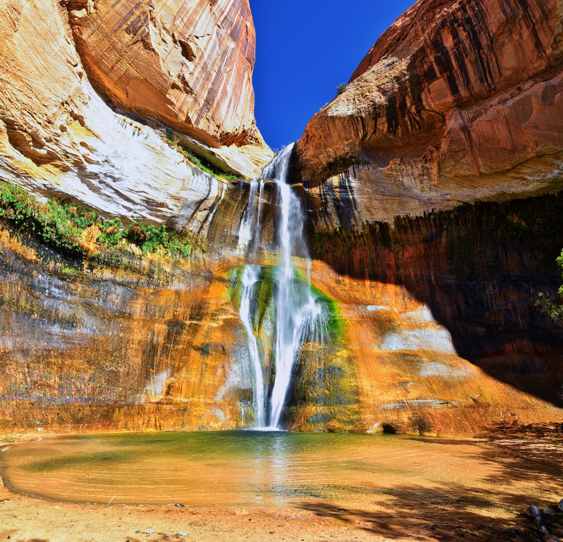 Waterfall cascading into a pool in a canyon with orange and brown rock walls and a blue sky.