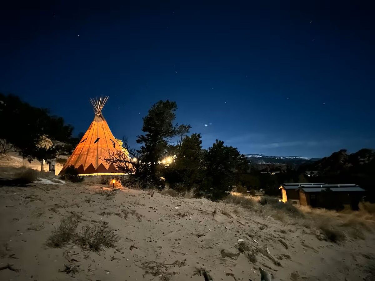 Night scene with illuminated teepee on a sandy hillside, trees and stars visible.