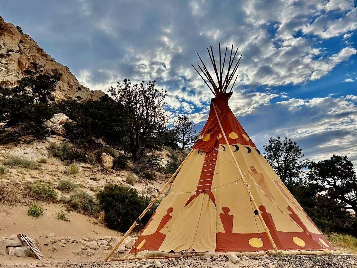 Teepee with painted symbols on a sandy hillside against a cloudy sky.