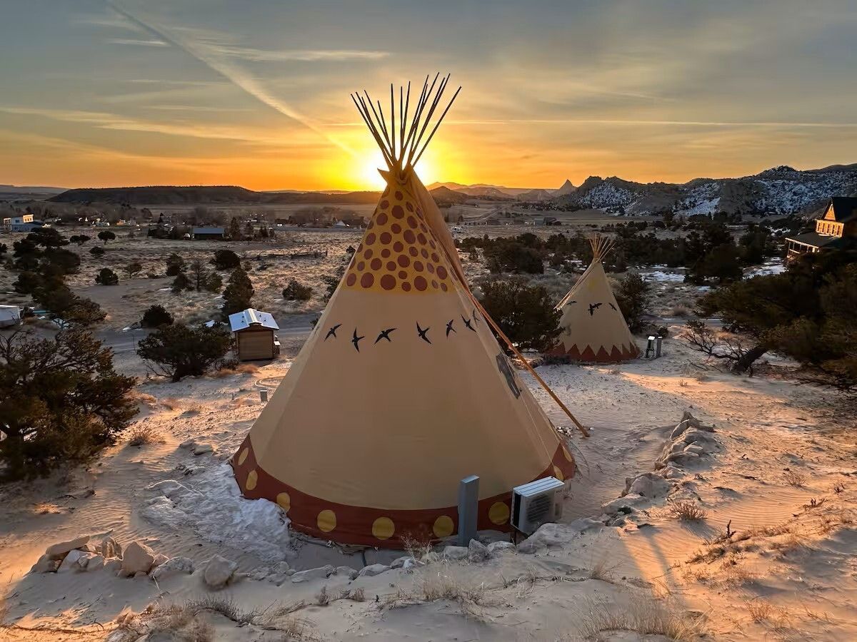 Two teepees in a desert landscape at sunset. Beige tents with decorative patterns; bright orange sun in background.
