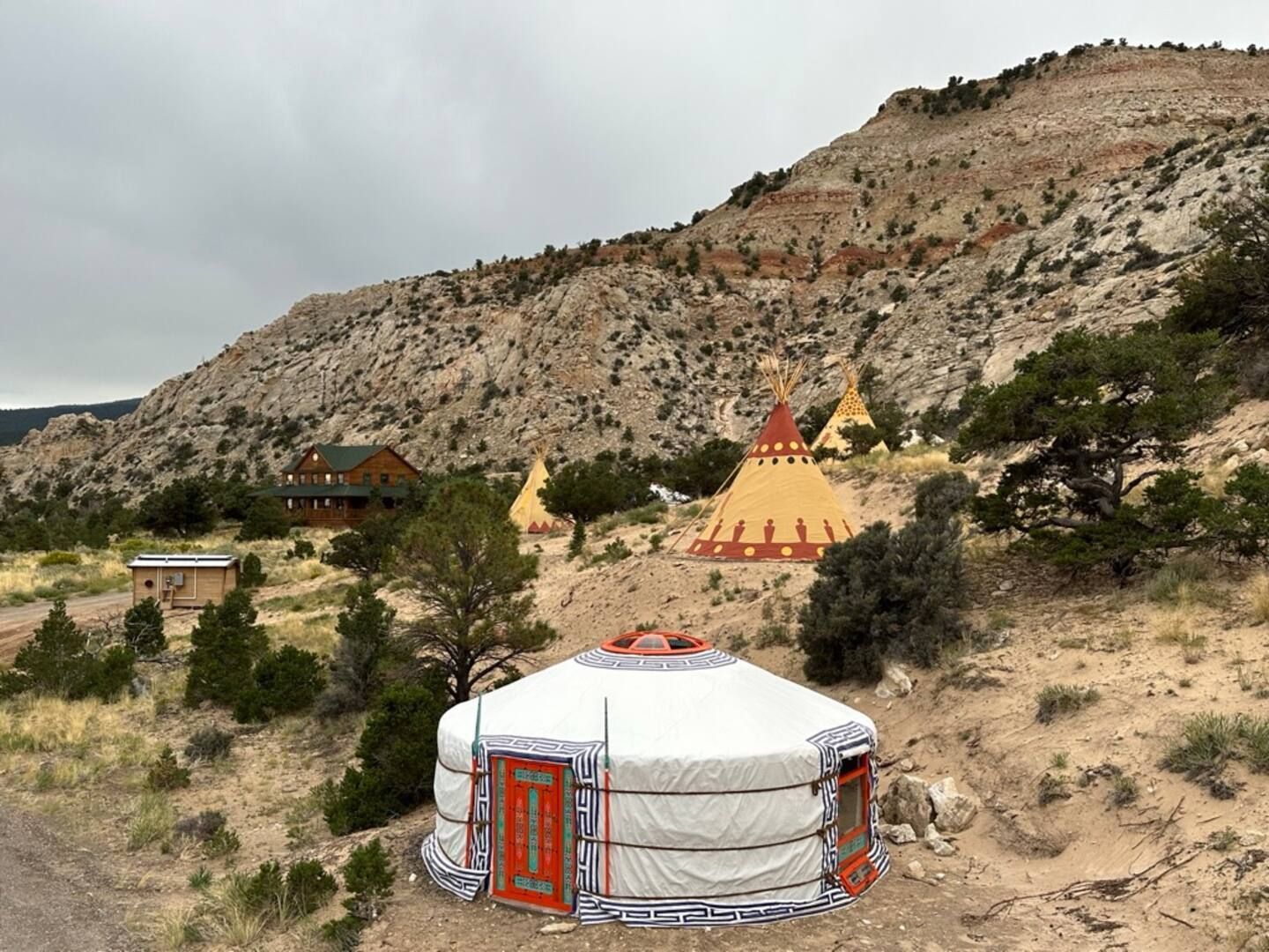 Yurt and tipis in a mountain setting. Brown and tan earth, overcast sky.