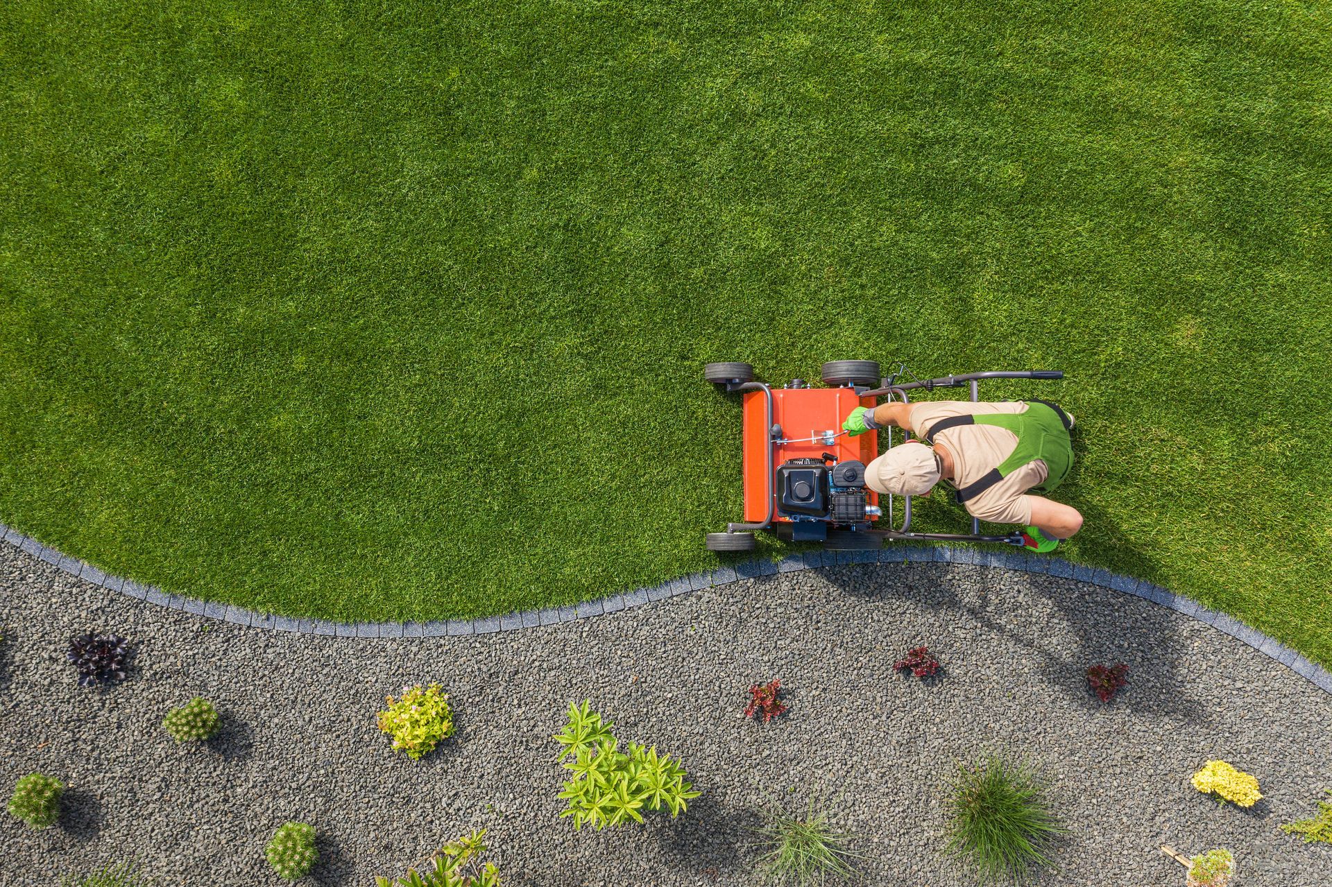 Overhead view: Person mowing a green lawn next to a gravel garden with plants.