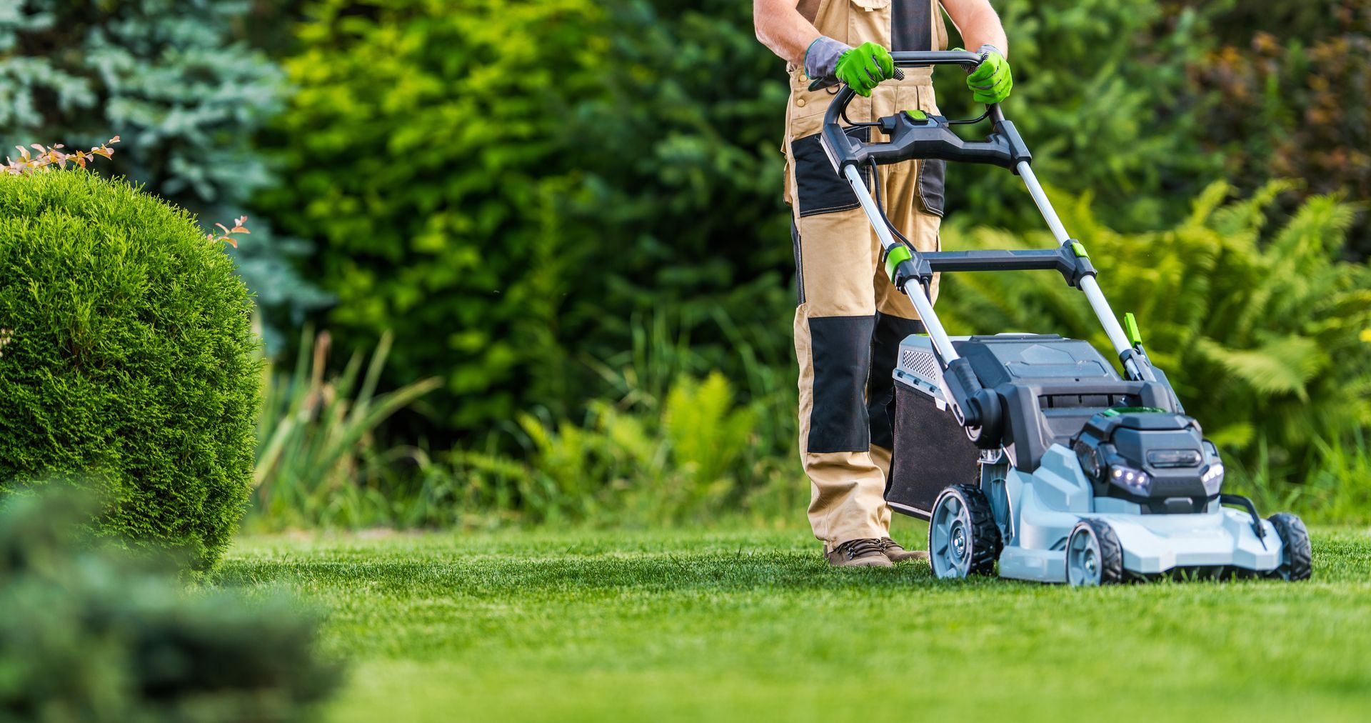 Person mowing a green lawn with a silver and black lawnmower. Green gloves. Lush green garden in the background.