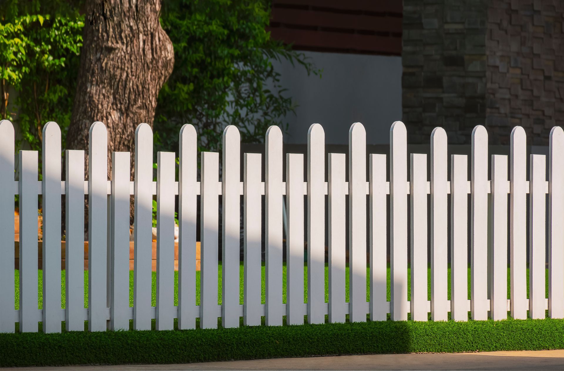 White picket fence in front of green grass, tree, and building.