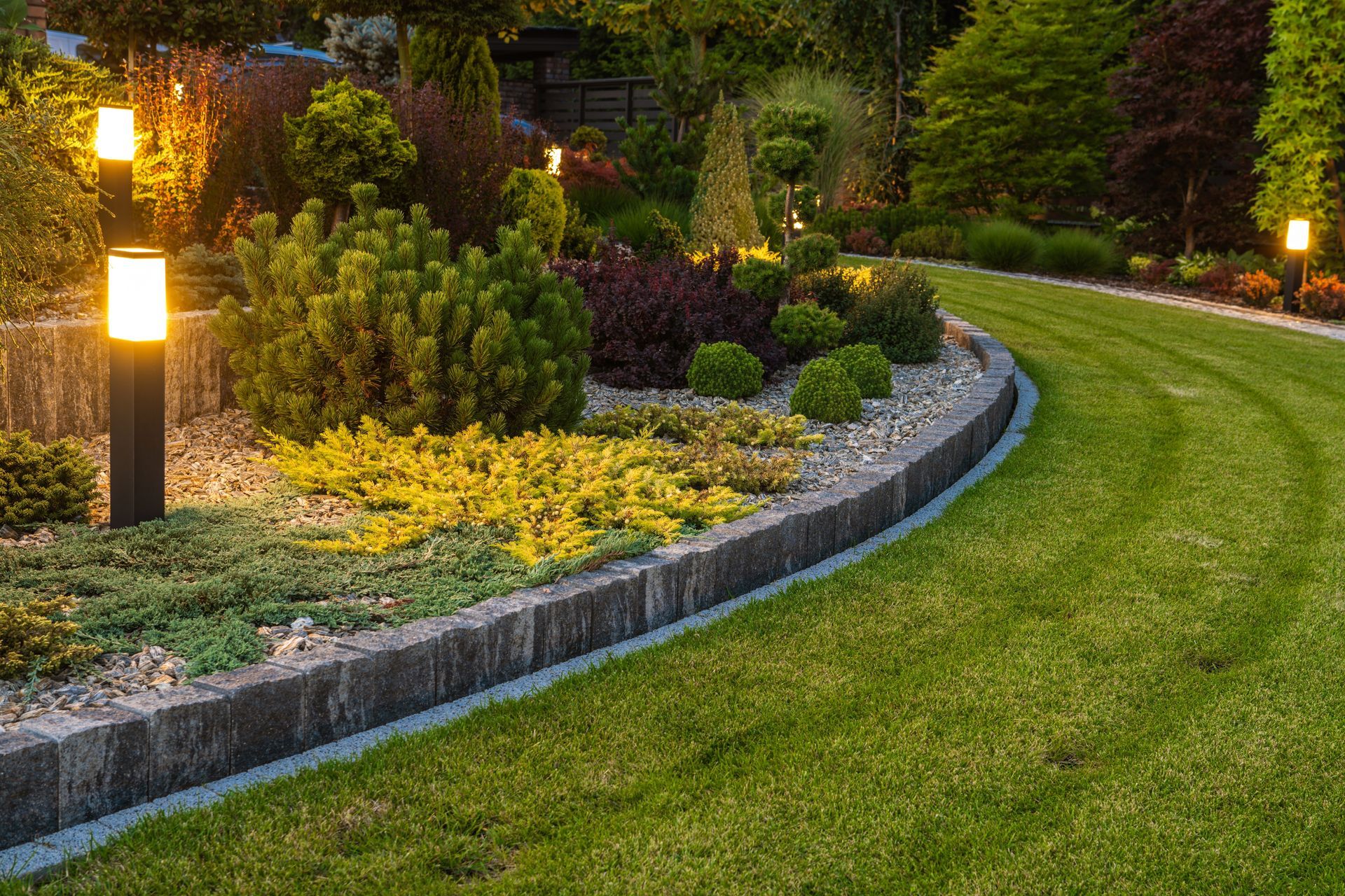 Garden bed with vibrant plants, illuminated by outdoor lights, alongside a green lawn.