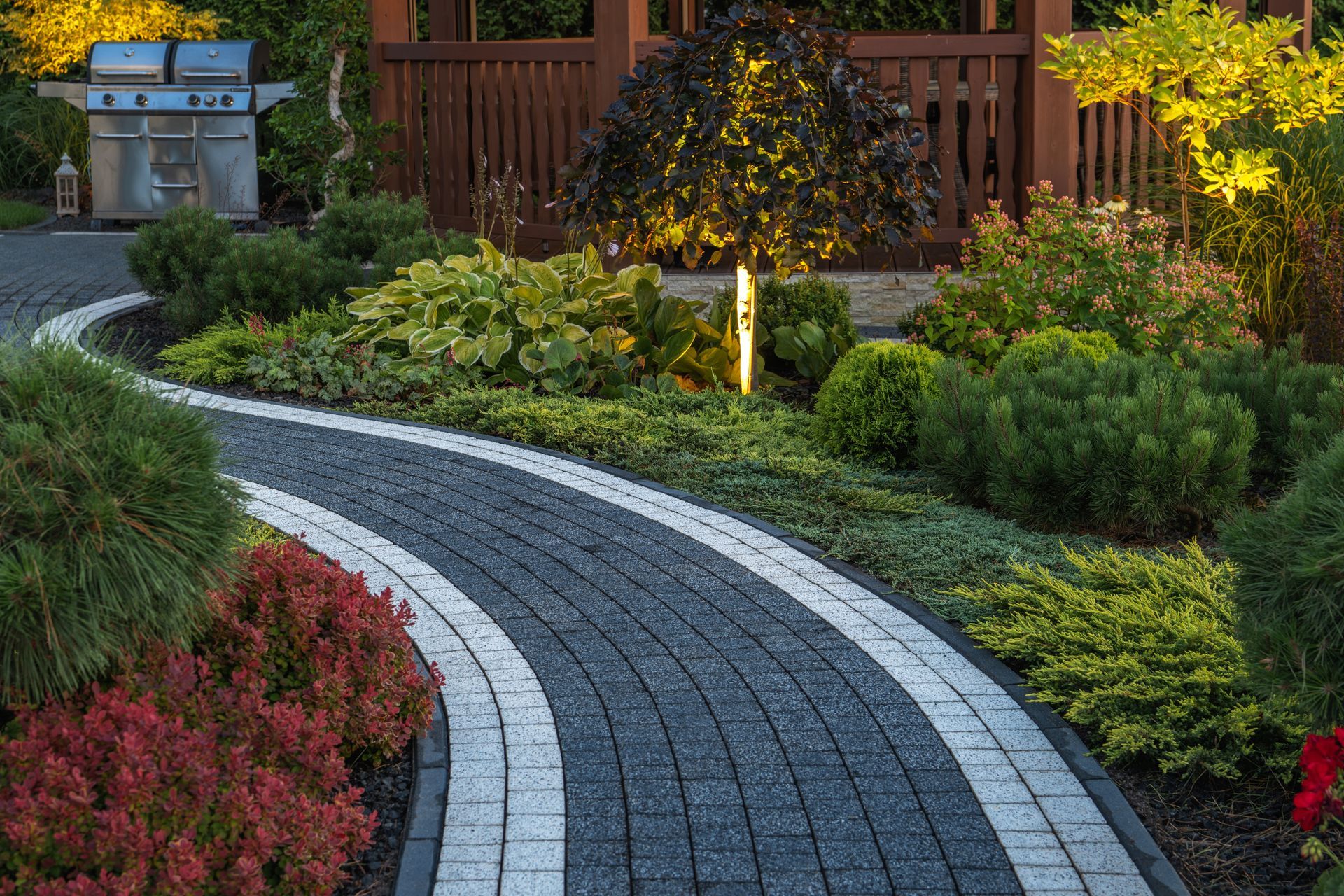 Curving brick pathway through a lush garden with bushes, grill, and gazebo.