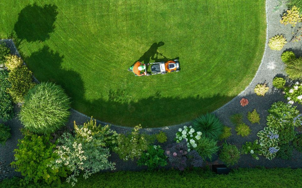 Aerial view of a person mowing a curved lawn surrounded by a rock path and various plants, creating a circular design.