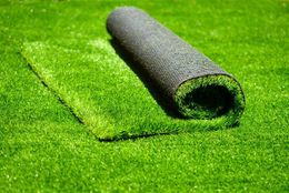 Roll of artificial turf unrolled onto a patch of green turf, showing a close up of the blades and backing.