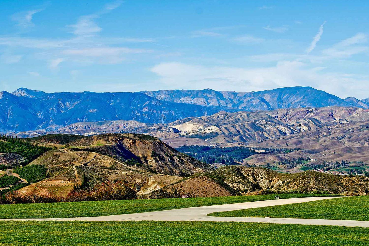 A dirt road winds through a grassy field with mountains in the background.