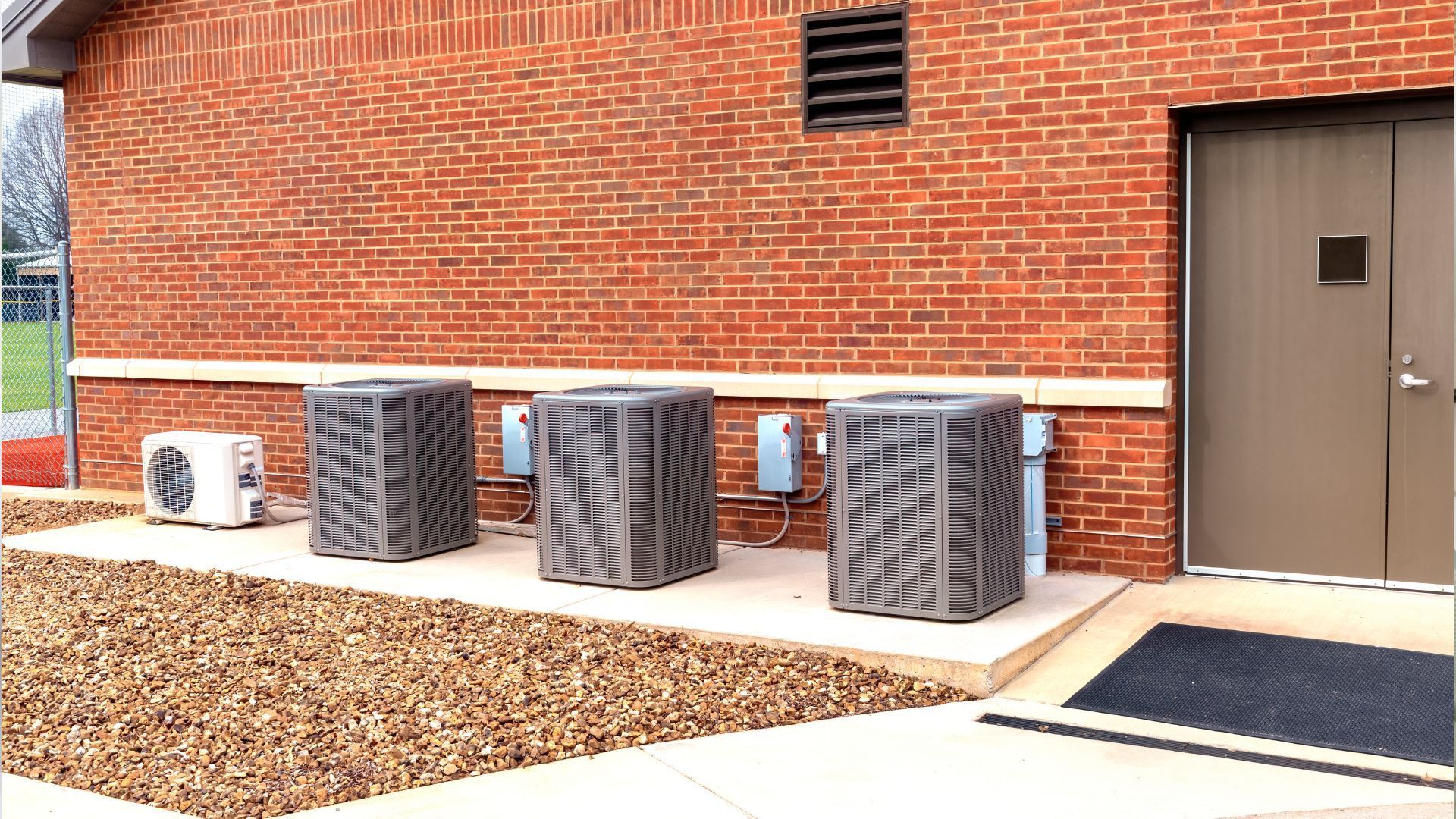 Brick building with air conditioning units, brown gravel, and a gray door.