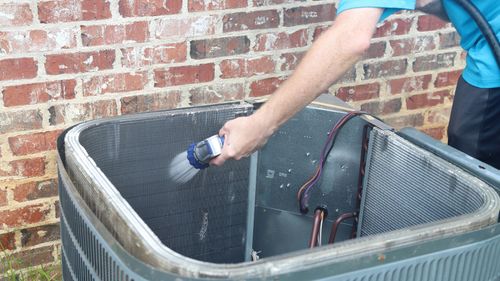 Person cleaning an AC unit with a brush and hose near a brick wall.